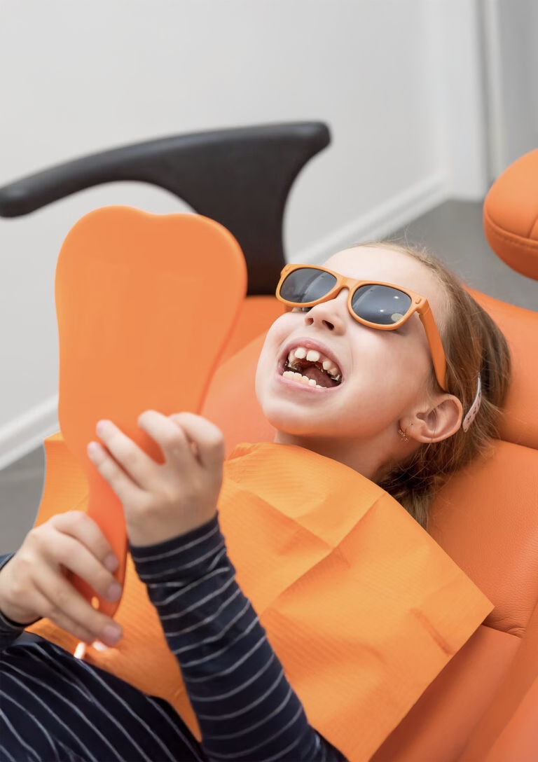Young Girl Wearing Braces And Sunglasses Looking At Herself In Mirror At Orthodontic Office